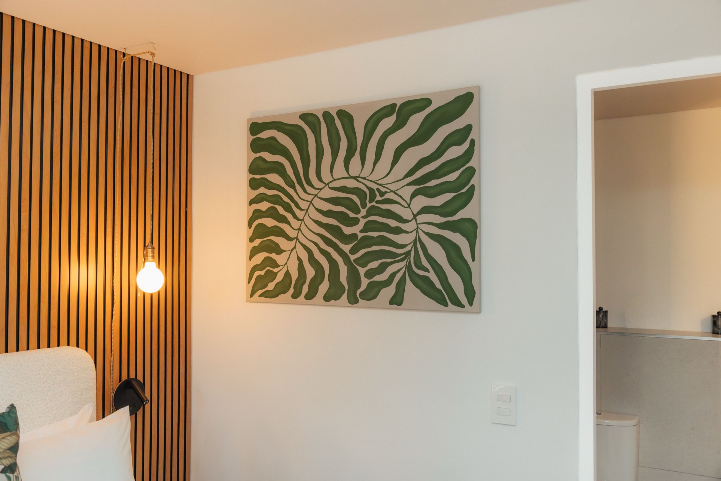 The green leaf canvas on the bedroom wall lit by a pendant bulb, with the bathroom doorway beyond
