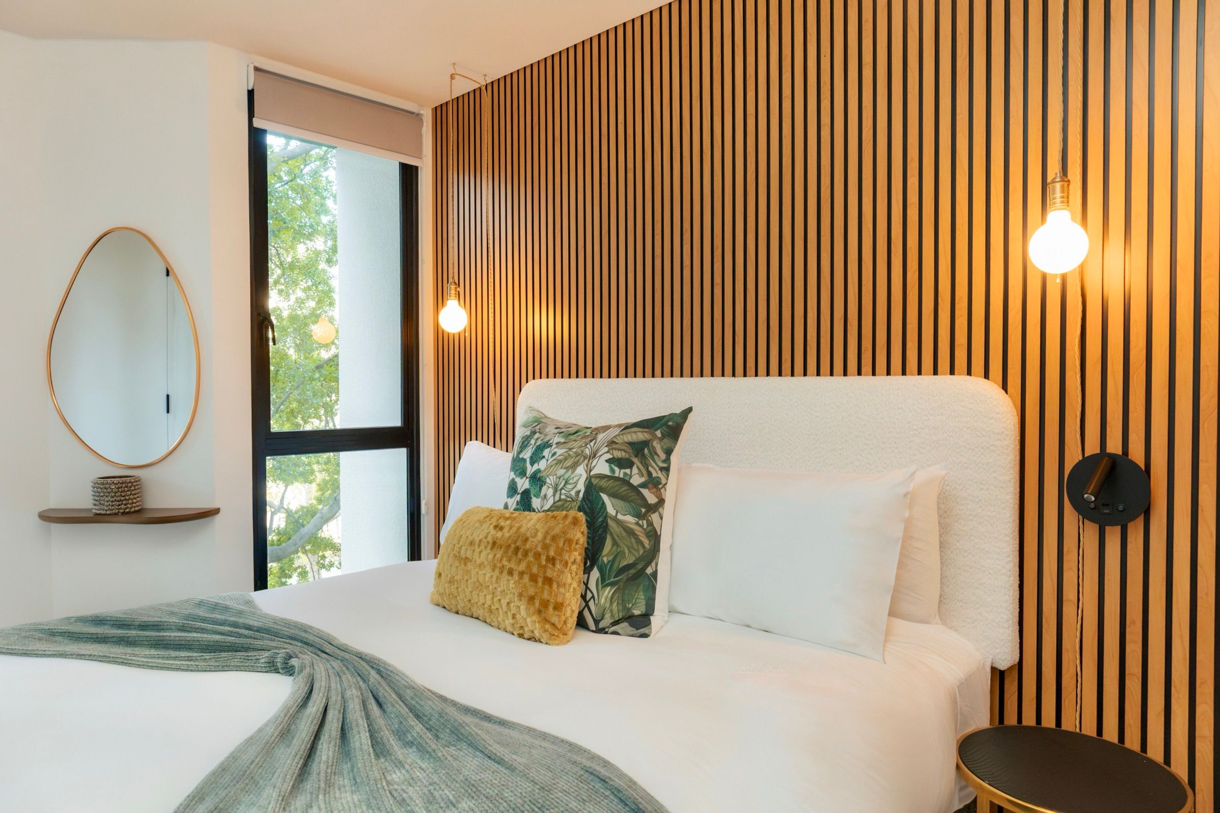 The bed against the slatted oak wall, with the tree visible through the bedroom window and a brass oval mirror on the left