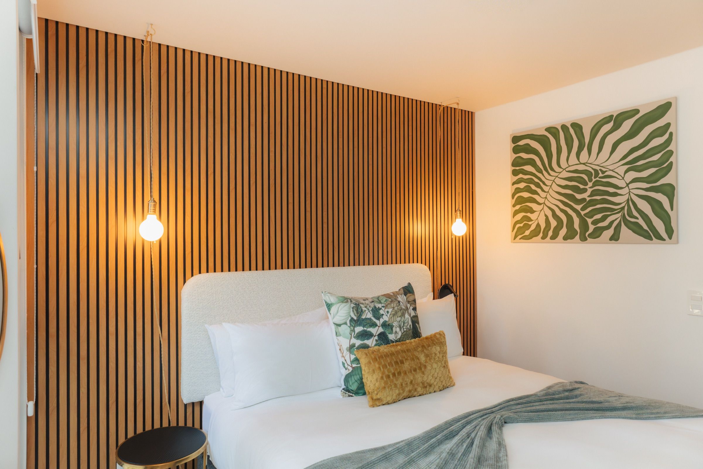 The bedroom with its slatted oak feature wall, two hanging pendant bulbs, white linen bedding and a green leaf canvas above