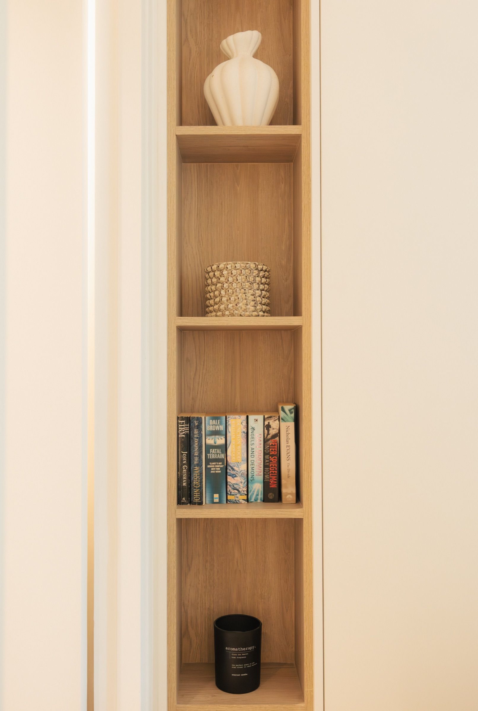 A built-in oak shelving niche holding a ribbed ceramic vase, a stack of paperbacks and a candle