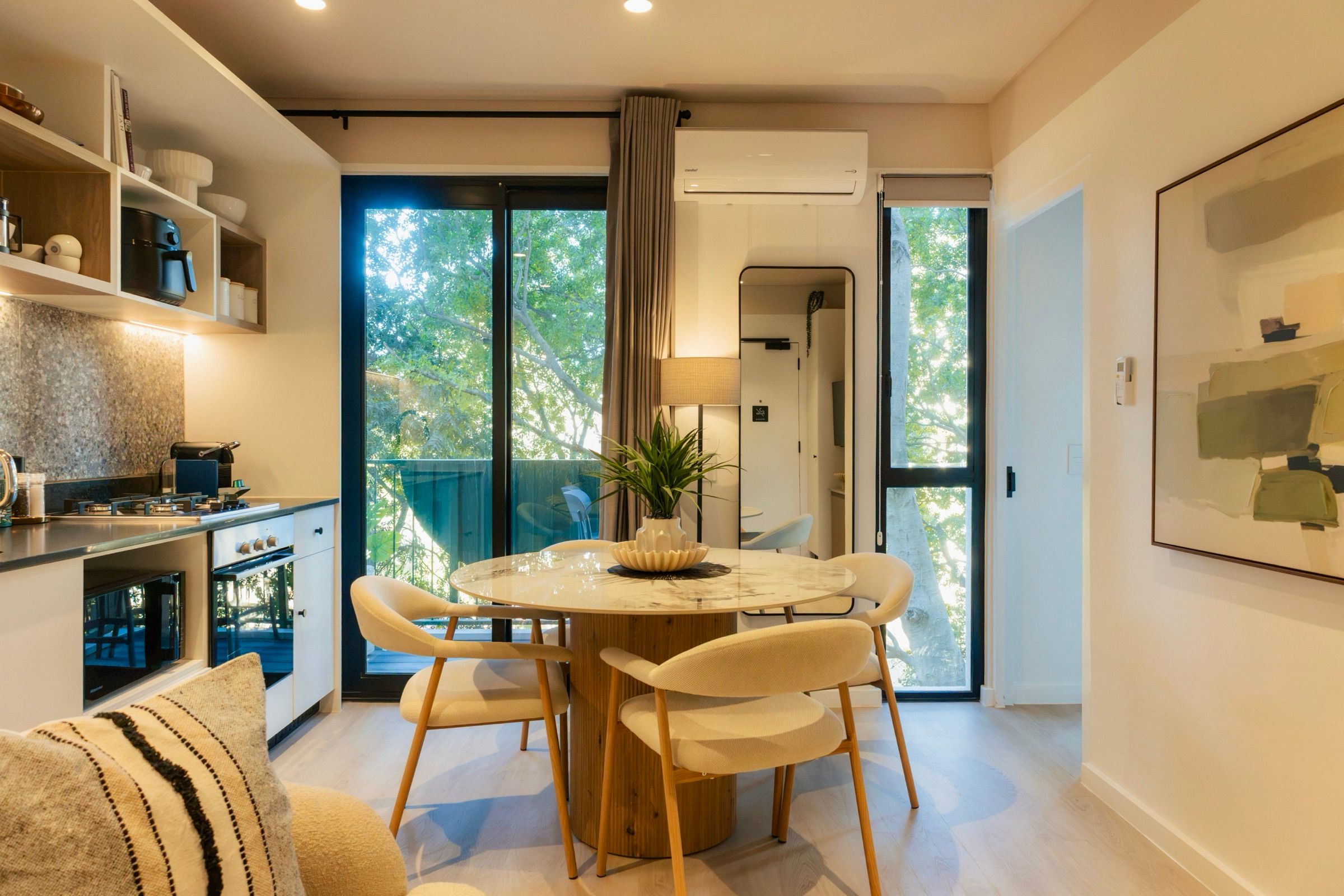 The kitchen and dining area together, with the tree canopy visible through the balcony doors and a framed green abstract on the far wall