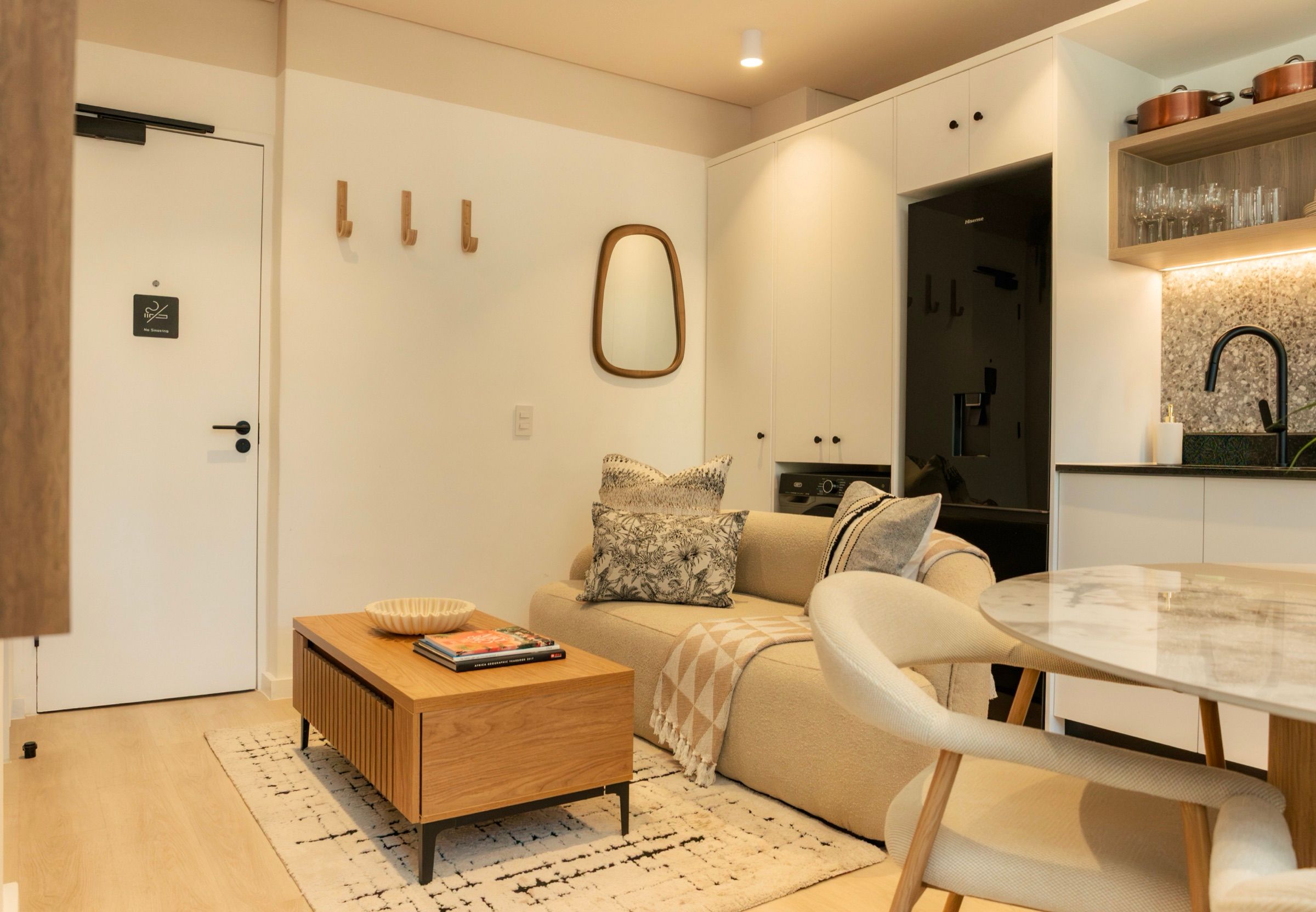 The apartment seen from the entrance: coat hooks and a rounded mirror, a cream sofa and oak coffee table on a berber rug, with the dining table and kitchen beyond