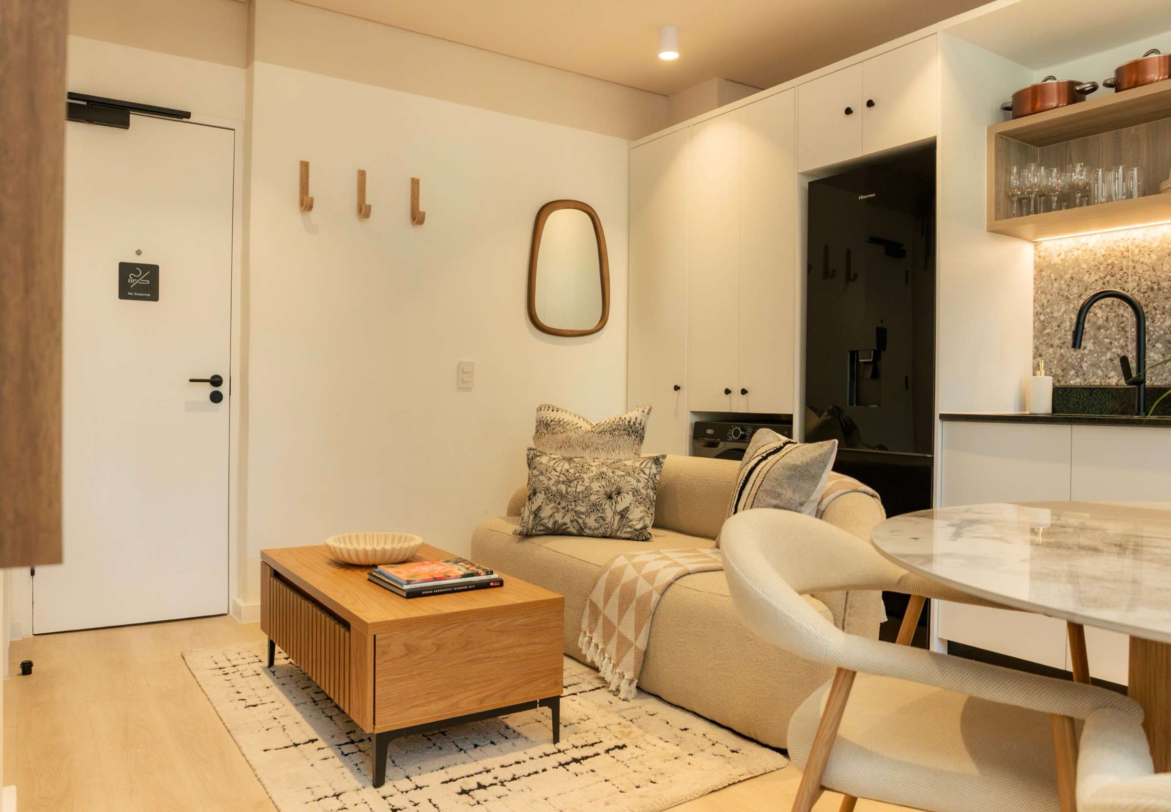 The apartment seen from the entrance: coat hooks and a rounded mirror, a cream sofa and oak coffee table on a berber rug, with the dining table and kitchen beyond
