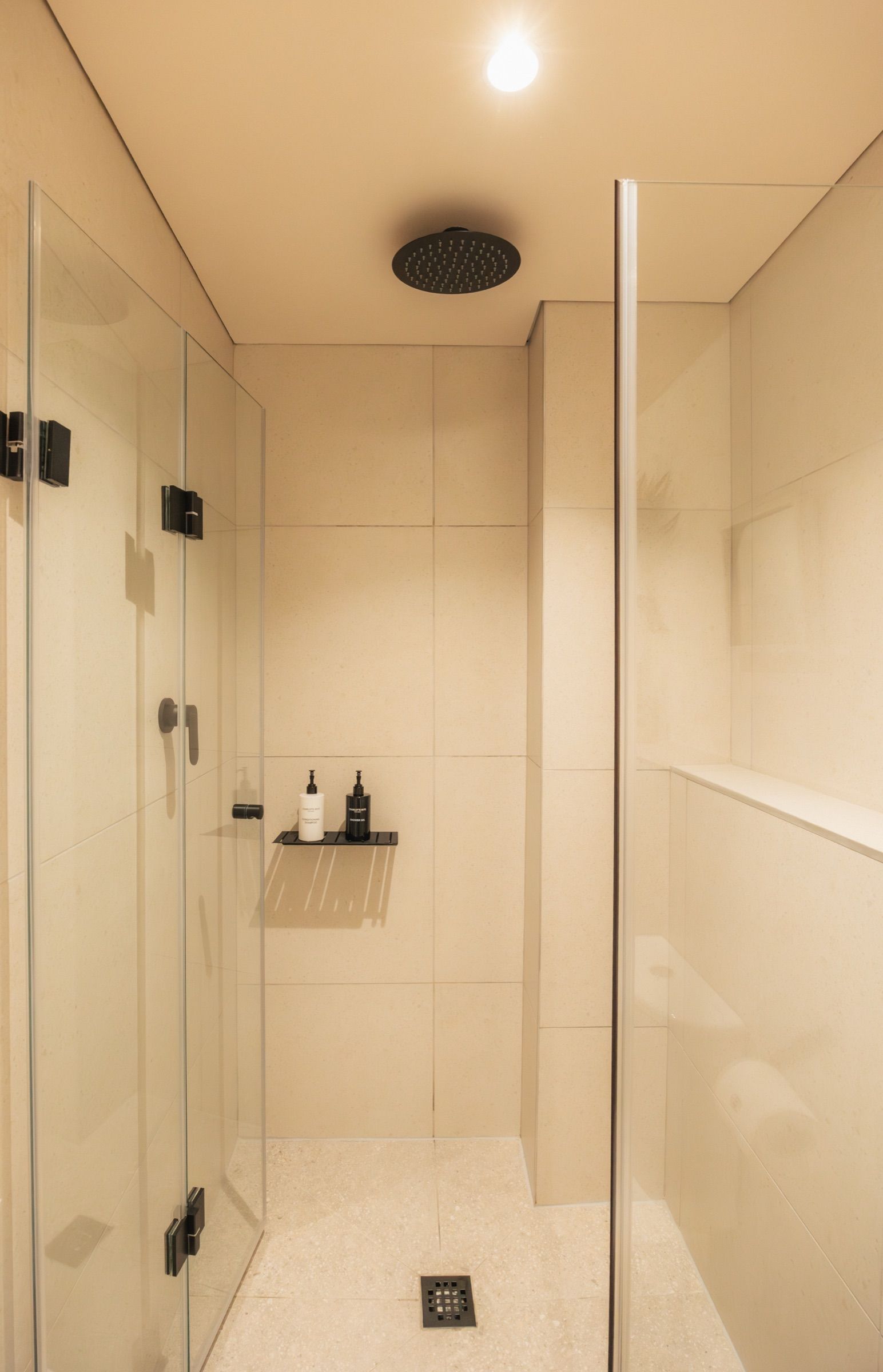 The walk-in shower with a black rainfall head, frameless glass and warm stone tiles